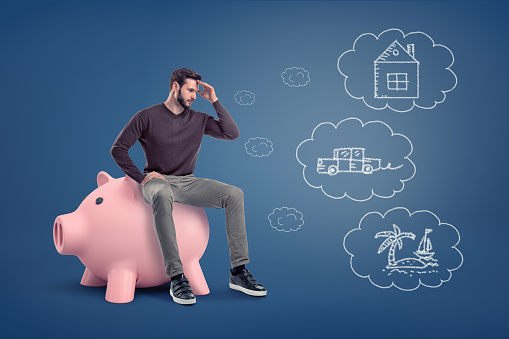 A thoughtful man sits on a large piggy bank near chalk drawings of a house, a car and an island. Choosing how to spend money . Savings and investments. Budget decisions.