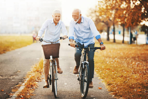 Couple of seniors enjoying a bike ride. Closeup front view of couple of cheerful seniors enjoying a bicycle ride in the city streets. It's mild October day with orange leaves and grass around them man 60s bicycle stock pictures, royalty-free photos & images