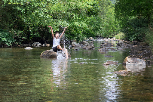 Teenage girl sitting on a rock, splashing water and having fun in a River Duddon in the Lake District National Park, UK
