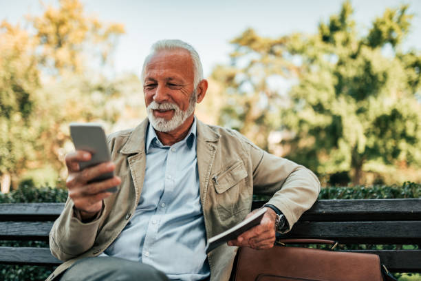 Senior man reading online news on smartphone outdoors. Senior man reading online news on smartphone outdoors. park bench photos stock pictures, royalty-free photos & images