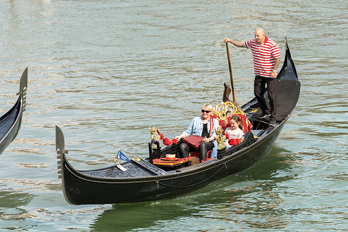 Venice, Italy, Sept 13, 2015: Two tourists sitting in the gondola with the gondolier, typical Venetian row boat. Sightseeing tour along the Grand Canal of the famous city. The gondola is a traditional, flat-bottomed Venetian rowing boat, well suited to the conditions of the Venetian lagoon. It is similar to a canoe, except it is narrower. It is propelled by a gondolier, who uses a rowing oar, which is not fastened to the hull, in a sculling manner and acts as the rudder.