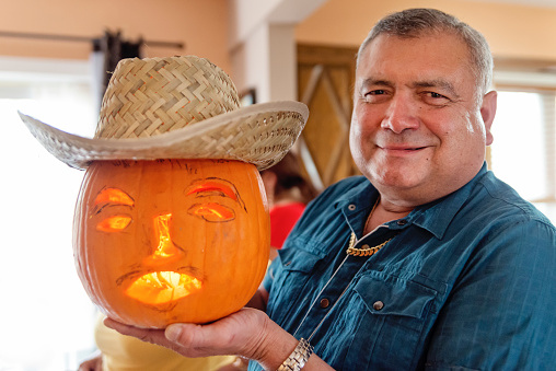 Proud senior Latin American man showing his freshly done carved pumpkin with an hispanic touch. Straw hat on the top of the pumpkin. Man is looking at the camera with a smile. Horizontal indoors waist up shot with copy space.