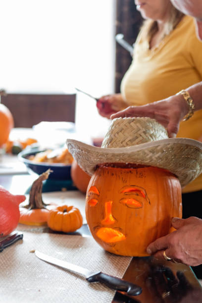 Latin American man carving and decorating pumpkin for Halloween. He is standing in the family kitchen and is putting a traditional straw hat on the finished pumpkin. Cropped view of his hands. No face. Vertical indoors waist up shot with copy space.