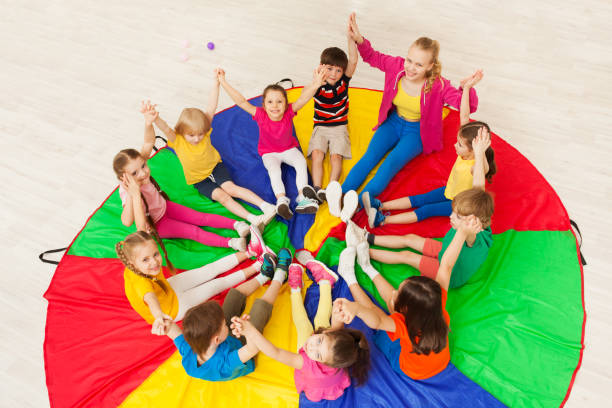Kids holding hands together with teacher in gym Top view portrait of young nursery teacher playing circle games with preschool children, holding hands together, sitting on rainbow parachute animator photos stock pictures, royalty-free photos & images