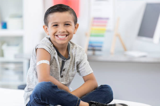 Little boy smiles proudly at camera after vaccination An adorable little elementary age boy sits cross legged on an exam table in his pediatrician's office.  He displays an arm bandage as he smiles for the camera. adhesive bandage photos stock pictures, royalty-free photos & images