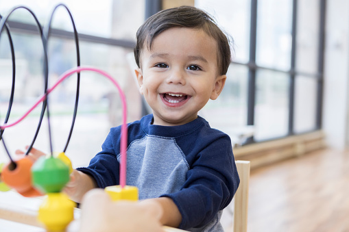 A happy toddler boy sits at a table in a doctor's waiting room and smiles for the camera.  There is a toy bead maze on the table.