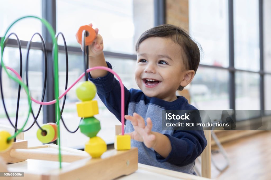 Toddler boy enjoys playing with toys in waiting room An adorable toddler boy sits at a table in a doctor's waiting room and reaches up cheerfully to play with a toy bead maze. Child Stock Photo