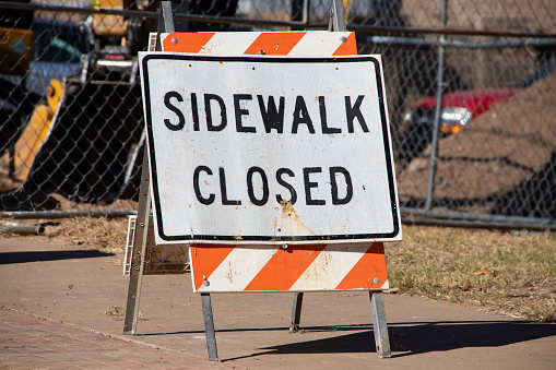 Sidewalk Closed standing sign on sidewalk in front of fence and construction equipment blurred in background