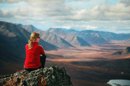 Blond woman in a red shirt perched atop a rock outcrop overlooking the majestic mountain-scape of the Tombstone Territorial Park in Yukon, Canada