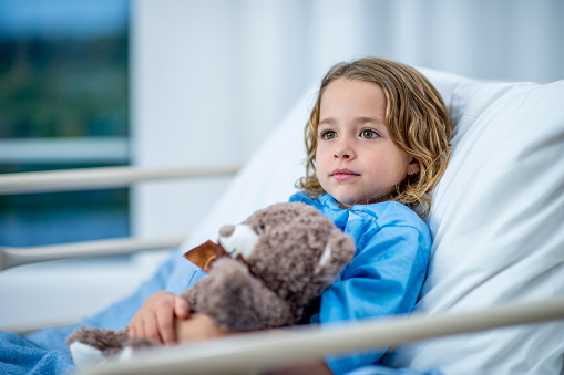 A girl of elementary age lies in a hospital bed and looks across the room with a noble, innocent expression. She is holding a teddy bear in her arms and wearing blue hospital clothing.