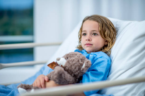 Young girl lying in a hospital bed looking stoic A girl of elementary age lies in a hospital bed and looks across the room with a noble, innocent expression. She is holding a teddy bear in her arms and wearing blue hospital clothing. sick child hospital bed stock pictures, royalty-free photos & images