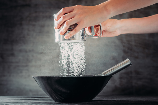 Woman sifting flour with sieve over black bowl on wooden table