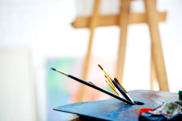 Close up of palette with brush at bright art studio. Wooden easel standing behind equipment on blurry background. Artist workspace in painter studio. Concept of hobby and talented people.