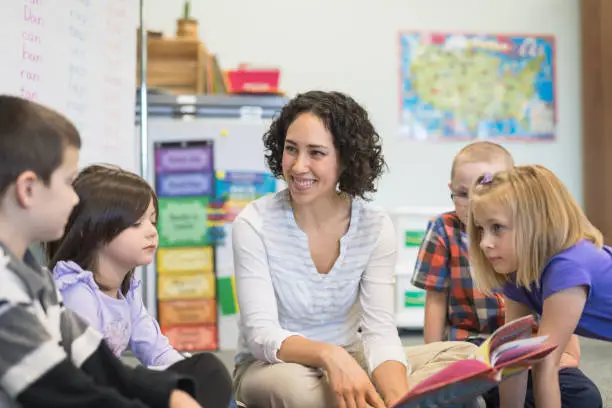 A young ethnic teacher reads a storybook to a group of first graders. They are gathered around her and listening intently as they sit on the floor.