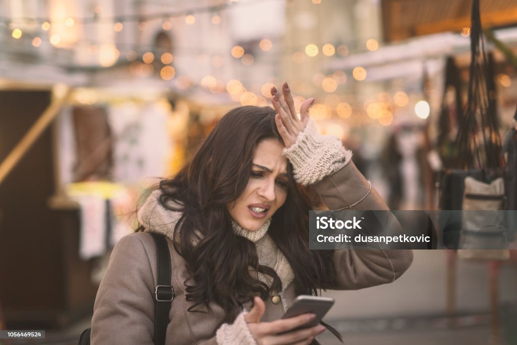 Young Caucasian woman using smart phone and holding her head while standing on the street. Reminder Stock Photo