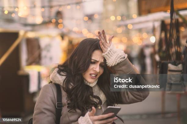Young Caucasian Woman Using Smart Phone And Holding Her Head While Standing On The Street Stock Photo - Download Image Now