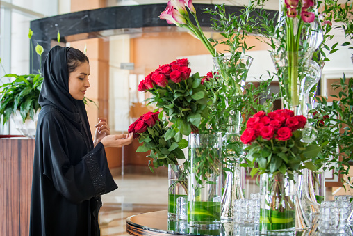 A young Emirati/Arab/Middle Eastern woman wearing a black abaya and hijab and surrounded by and potted plants and bouquets of long stemmed red roses and calla lilies arranged in vases, smiles while touching a rose in a florist shop.  Dubai, U.A.E., Middle East