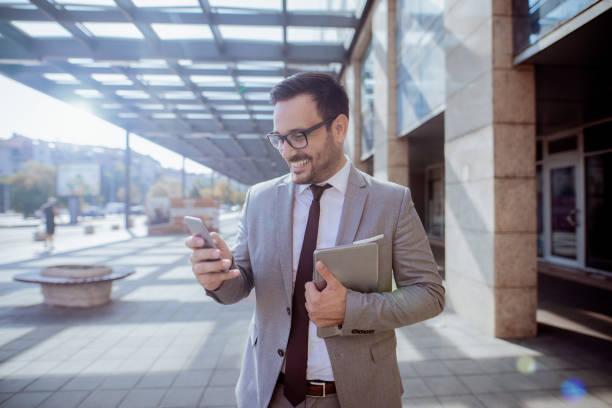 hombre de negocios con teléfonos inteligentes para leer/escribir mensaje de texto mientras está parado en frente de centro de negocios. en otros tablet de la mano. - street directory fotografías e imágenes de stock