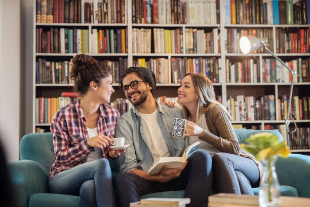 istock Two female students drinking coffee and male student holding book while sitting on sofa in library. 1056476898