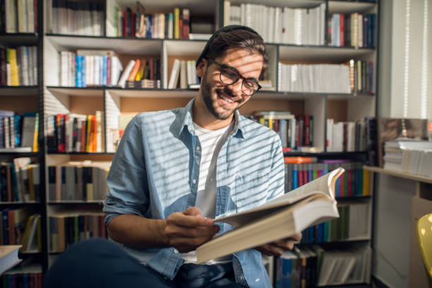 istock Happy male student sitting in library and reading book. In background bookshelf. 1056477304