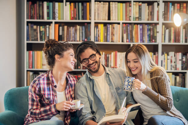 istock Happy students sitting on sofa and discussing book. In background bookshelf. 1056477510