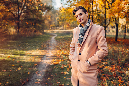 Young handsome businessman walking in autumn forest at sunset. Stylish guy wearing classic clothes and accessories. Fall fashion