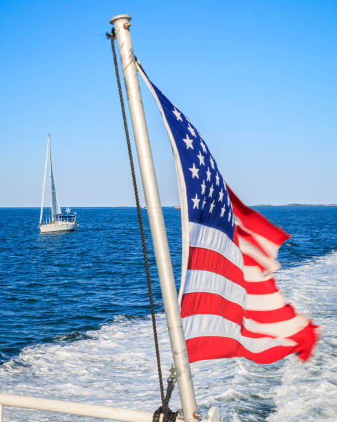 An American flag whips behind a speed boat creating a white wake on blue water. A sail boat is behind and to the right of the speed boat. A blue sky is the background.