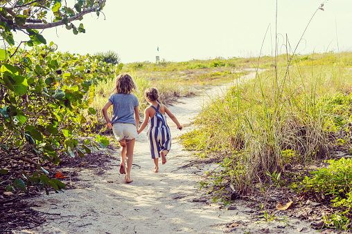 Two cute children, sisters or friends , barefoot, holding hands in a sandy tropical location, idyllic paradise, sand dunes, they run hand in hand, in a rush on their way to the beach. Heartwarming moment of childhood. Age 5 and 7