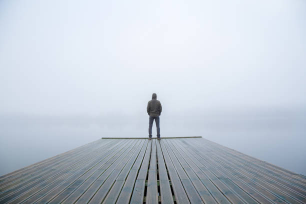 Young man standing alone on edge of footbridge and staring at lake. Mist over water. Foggy air. Early chilly morning in autumn. Beautiful freedom moment and peaceful atmosphere in nature. Back view. Young man standing alone on edge of footbridge and staring at lake. Mist over water. Foggy air. Early chilly morning in autumn. Beautiful freedom moment and peaceful atmosphere in nature. Back view. man regret stock pictures, royalty-free photos & images