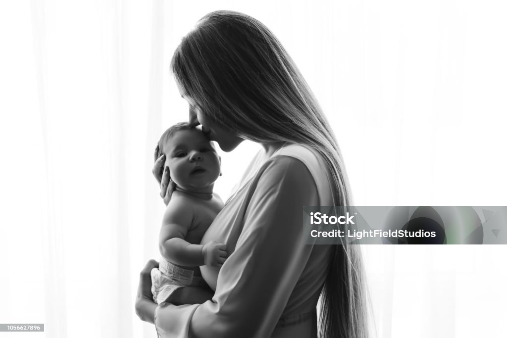 photo noir et blanc de mère embrassant le petit garçon de bébé en face de rideaux à la maison - Photo de Mère libre de droits