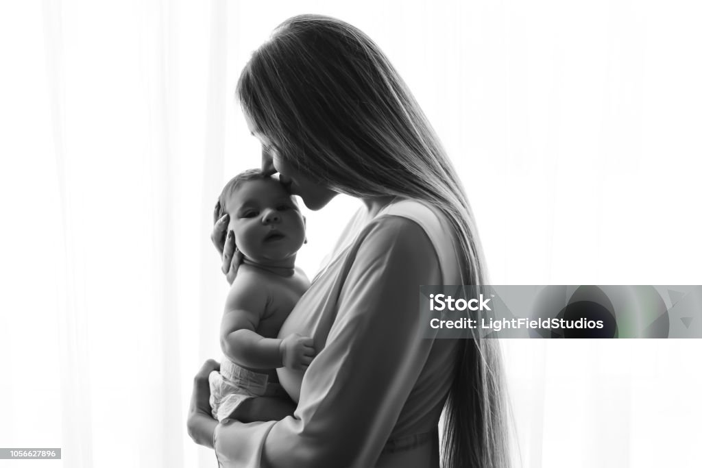 foto preto e branca da mãe beijando o menino na frente de cortinas em casa - Foto de stock de Mãe royalty-free