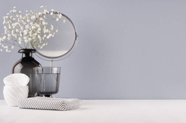 モダンなエレガントな女性ドレッシング テーブル花、ミラー、グレー色の壁と白い木の板に銀化粧品袋と黒のガラスの花瓶。