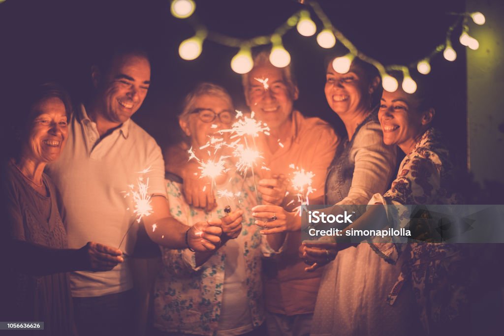 group of caucasian people friends with different ages celebrate together a birthday or new year eve by night outdoor at home. lights and sparkles  with cheerful women and men having fun in friendship Senior Adult Stock Photo