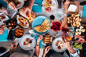 Above aerial view of group of friends having fun eating together at lunch or dinner with a table full of different and colorful food and technology mobile phone. mix of hands of caucasian people taking somethings to eat together
