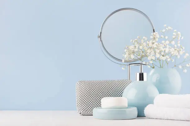 Photo of Bathroom decor for female in light soft blue color - circle mirror, silver cosmrtic bag, white flowers, towel, soap and ceramic smooth vase on white wood table.