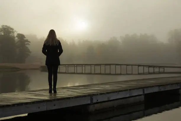 Photo of Young woman standing alone on lake footbridge and staring at sunrise in gray, cloudy sky. Mist over water. Foggy air. Early chilly morning. Dark, scary moment and gloomy atmosphere. Back view.