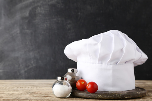 Chef hat with salt, pepper and tomato on wooden table