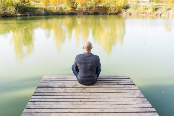 istock Young bald man in the lotus pose sits on the wood pier. Back view 1056665652