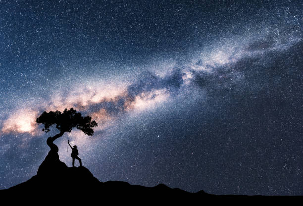 milky way and silhouette of woman under the tree growing from the rock on the mountain at night. space background with starry sky, beautiful galaxy and girl. scenery with milky way and people. travel - milky way galaxy space star imagens e fotografias de stock