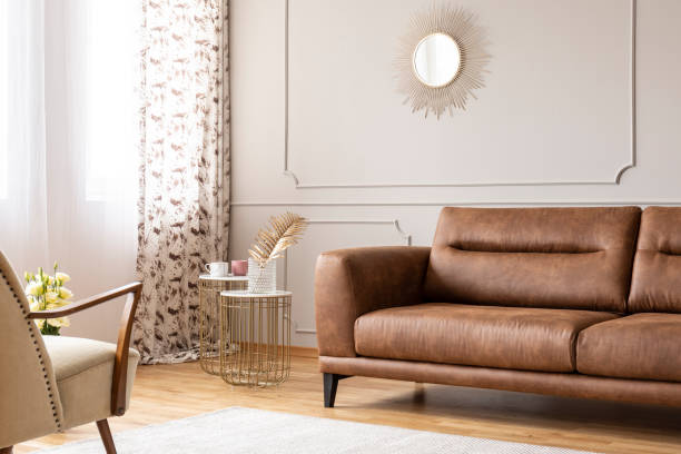 istock Gold mirror on grey wall with molding above leather settee in flat interior with tables and armchair. Real photo 1056681258