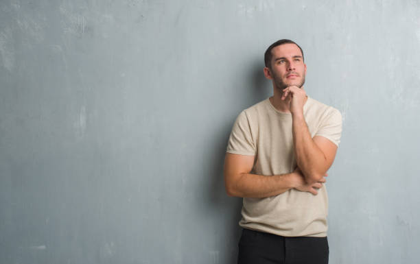 istock Young caucasian man over grey grunge wall with hand on chin thinking about question, pensive expression. Smiling with thoughtful face. Doubt concept. 1056688126