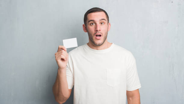 istock Young caucasian man over grey grunge wall showing blank visit card scared in shock with a surprise face, afraid and excited with fear expression 1056689584