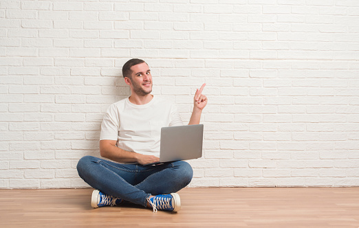 Young caucasian man sitting over white brick wall using computer laptop very happy pointing with hand and finger to the side
