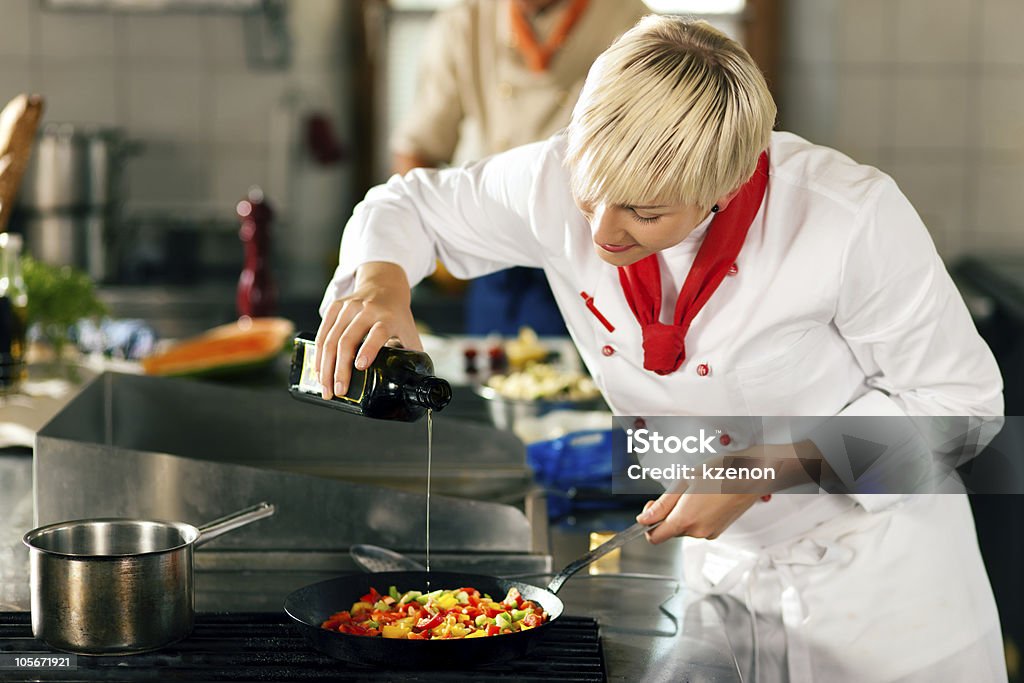 Chefs in a restaurant or hotel kitchen cooking Two chefs in teamwork - man and woman - in a restaurant or hotel kitchen cooking delicious food, she is putting olive oil in the ratatouille Chef Stock Photo