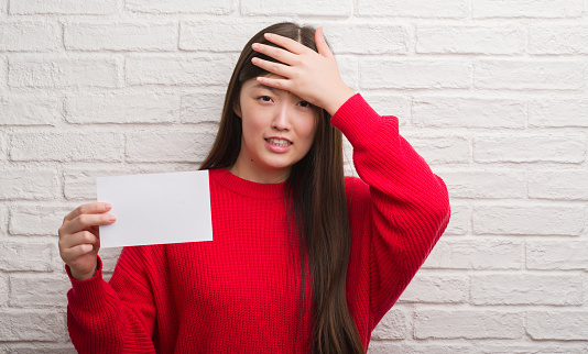 Young Chinese woman over brick wall holding blank paper stressed with hand on head, shocked with shame and surprise face, angry and frustrated. Fear and upset for mistake.