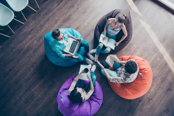 vue grand angle haut de diversité hipster moderne et élégant jeune homme assis dans les fauteuils colorés sur les travaux de plancher de bois ensemble sur le sac de chaise de tâche sit ses camarades de classe - coworking photos et images de collection