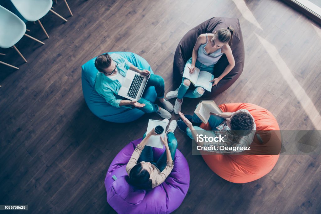 Alta vista dall'alto angolo della diversità elegante e moderno hipster giovane sedersi su poltrone colorate sul pavimento in legno lavorare insieme sul compito sit borsa sedia compagni di classe - Foto stock royalty-free di Nuova impresa