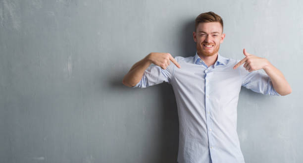 istock Young redhead business man over grey grunge wall looking confident with smile on face, pointing oneself with fingers proud and happy. 1056761400