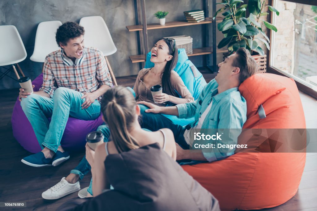 Rejoice and cheerful trandy youths sitting in a loft style room with coffee in hands tell funny stories and humorous joke to each other and aloud laugh without restraint with big toothy smile Coworking Stock Photo