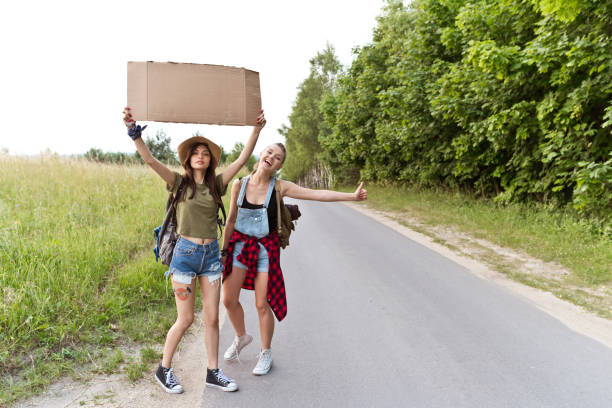Two hipster girls hitchhiking together Outdoor shot of two young women standing on the road and hitchhiking. hitchhiking stock pictures, royalty-free photos & images
